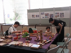 長机に敷かれた紫色の布の上に、木でできた様々な列車や家のおもちゃが並べられ、大人と子供が一緒に遊んでいる様子の写真