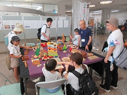 長机に敷かれた紫色の布の上に、木でできた様々な列車や家のおもちゃが並べられ、親子連れやスタッフが囲んで遊んでいる様子の写真