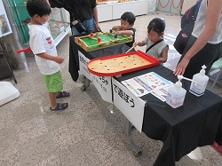 黒い布が敷かれた長机に木でできた様々なおもちゃがあり、机を囲んで複数の子供たち遊んでいる様子の写真