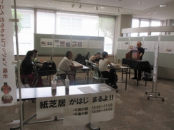 「紙芝居がはじまるよ！！」と書かれた紙に長机に貼られ、その奥で椅子に座って紙芝居を見ている複数の観客と、それを演じている坂井謙さんの写真