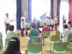 明るいホールのような会場で、合唱服のような制服を着た9人ほどの児童が合唱しており、ピアノが置かれた傍で指揮者らしき大人が立っている前で、数人の参加者が着席して楽譜を持って聞いている写真