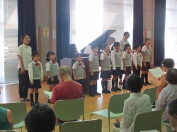 育館かホールのような場所で、合唱服のような制服を着た数人の小学生たちが、中央に置かれたピアノと指揮者らしき大人の前で、着席した聴衆に向かって歌っている様子の写真