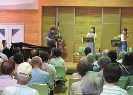 男女4名がピアノ、クラリネット、バイオリン、コントラバスをステージ上で演奏し、その様子を多くの観客が椅子に座って鑑賞している写真