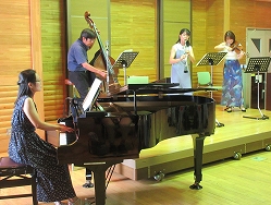 男女4人の演奏家がステージ上でピアノ、コントラバス、クラリネット、バイオリンを演奏している様子の写真