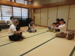 和室の畳の上で、数人の女性が小さな子ども膝に乗せ座り、和やかな雰囲気の中で手遊びをしている写真