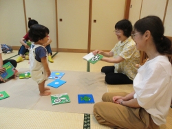 和室で、畳の上に数冊の絵本が広げられており、大人2人が座って立っている小さな子どもに向き合い、1人の大人が子どもに絵本を見せている様子の写真