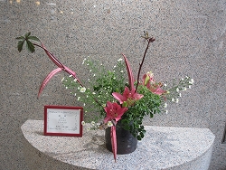 濃いピンク色のユリのような花と細長い赤い茎、白い小花や緑の葉を組み合わせた華やかな生け花の作品と赤い額縁のプレートが写っている写真