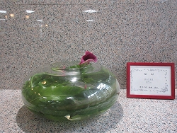 透明な丸いガラスの花器に濃い緑色の葉と濃い紫色のカラーのような花が生けられており、右側には赤い額縁のプレートが添えられた生け花の作品写真