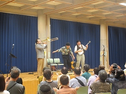 ステージ上で、トロンボーン、バンジョーを演奏する男性と、タッブシューズを履きステップを踏んでいる様子の三人組のジャズバンドのコンサート風景を、聴衆の後方から捉えた写真