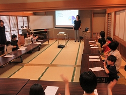 前方にスクリーンが写され右手に講師が立ち。和室の畳の上に長机をロの字型に配置した周りに座る複数の参加者が手を動かし手話の勉強会をしている様子の写真