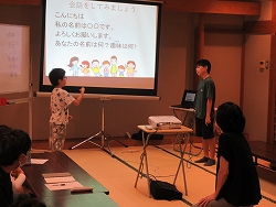 和室の会場で、スクリーンに投影された「会話をしてみましょう」という文字と前方に向かい合って立つ2人の男の子が手話で話をしている様子の写真