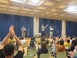 ステージ上で、三人のミュージシャンが観客に向かって手を挙げ、大勢の観客が拍手している様子を、観客席の後方から捉えた、コンサートが盛り上がっている様子の写真