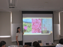 プレゼンテーションを行う女性がプロジェクタースクリーンに映し出された野外のバラ、集められた大量のバラ、液体が入ったガラス器具の3分割の画像の前で花について説明している様子の写真