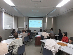 会議室のような部屋で、一人の女性がプロジェクターに映し出されたスライドを見せる形で、十数人の聴衆に向かってプレゼンテーションを行っている様子の写真