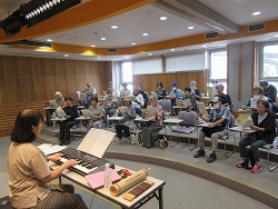 室内で、女性の指導者が電子キーボードを弾きながら、楽譜を持った多くのシニア世代の参加者たちに囲まれ、合唱をしている様子の写真