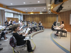 室内のひな壇の座席を備えた半円形の座席で、一人の女性講師が参加者のグループに向かって楽譜を使った合唱または音楽の授業を教えている様子の写真