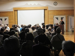 会場で、多くの聴衆が背中を向けて並び、演台に立つ一人の男性講師が正面の白いスクリーンに向かって講演を行っている様子の写真