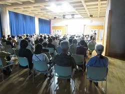 会場で、多数の聴衆が椅子に座り、遠くのステージ上で行われているイベントや講演を鑑賞している様子を後ろから写した写真