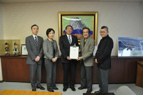 五人の男女が室内で横一列に立ち、中央の男性二人が白い書類の入った額縁のようなものを挟んで握手を交わしており、その背後には富士山の絵画が飾られた応接室での集合写