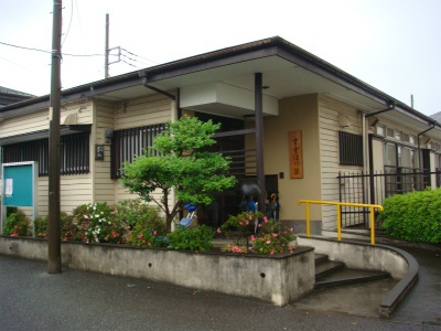 薄茶色の壁と黒い窓枠の平屋のすずはり荘の玄関前に、ツツジや低木が植えられた低い石垣があり、右側には黄色い手すりのついた緩やかなスロープが設けられている外観写真