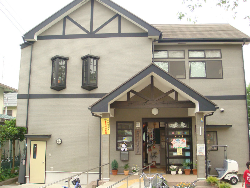 淡いグレーの壁と黒い木枠が特徴の洋風2階建ての建物で、正面の玄関前にはスロープがあり、軒先には様々な装飾や植木鉢が飾られている沼代児童館の外観写真