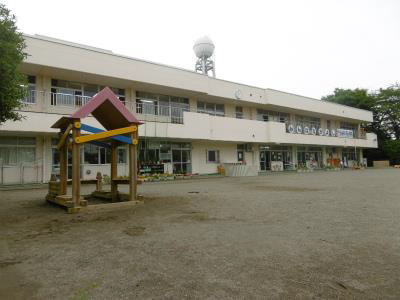 園庭の土の上に木製の遊具が置かれ、その後ろには白い2階建ての建物が広がり、屋上には給水塔のような鉄塔が見える幼稚園の外観写真