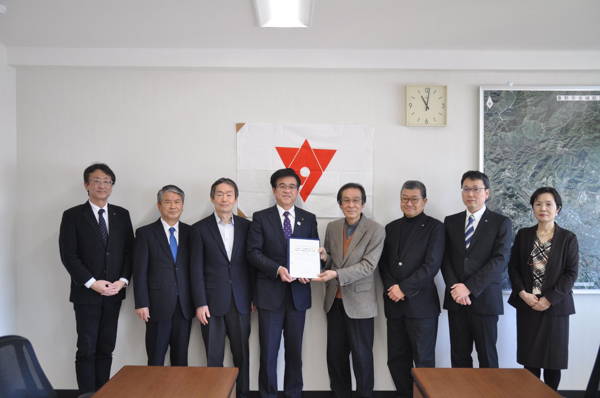 白と赤の旗を背景に、スーツ姿の7人の男性と1人の女性が、中央の市長と斉藤会長が報告書を持って横並び立っている集合写真