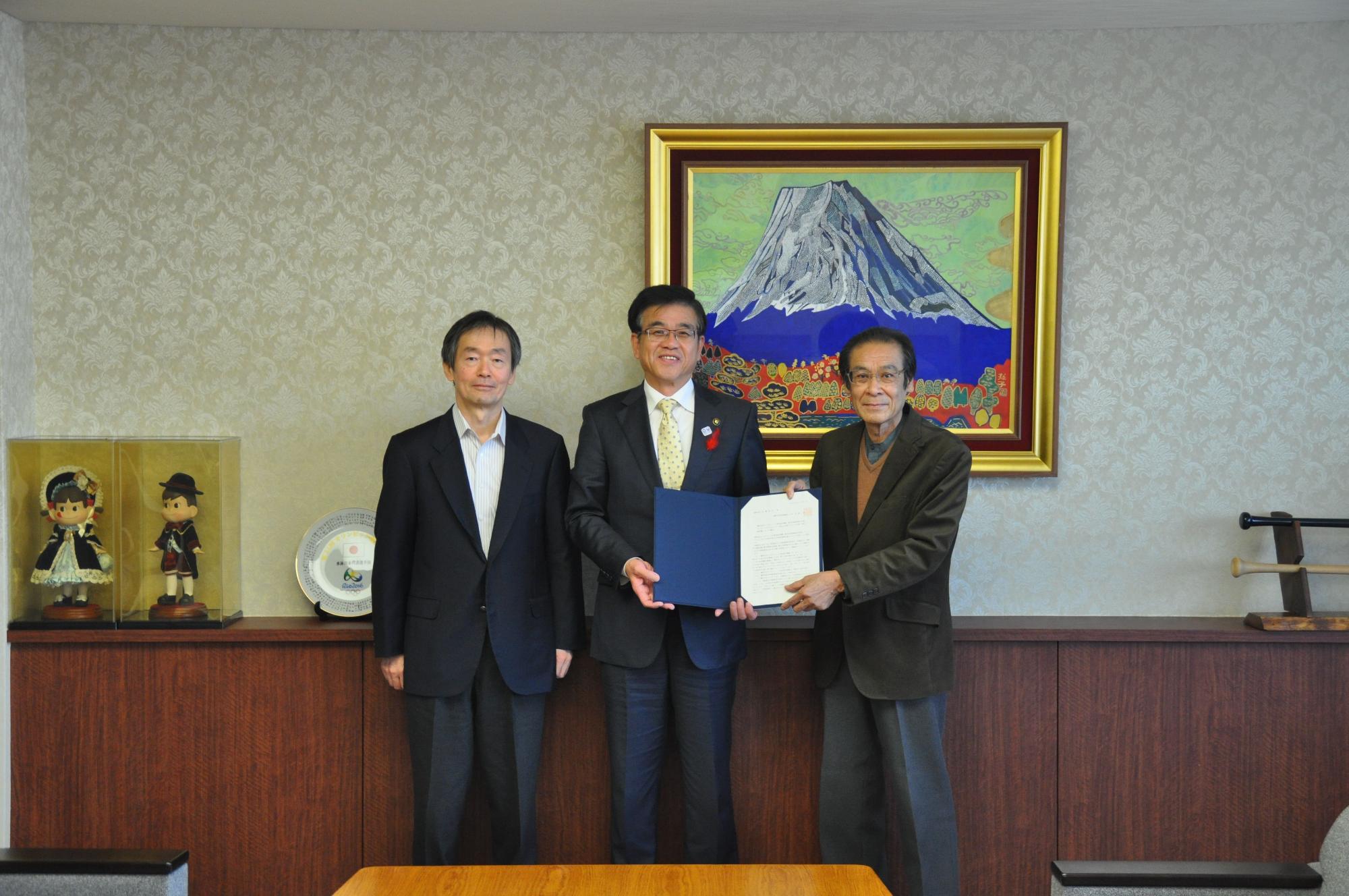 薄茶色の壁紙の部屋で、富士山の絵画と日本人形が飾られた棚を背景に、中央の市長と斉藤会長が一緒に報告書を持って並び立つ集合写真