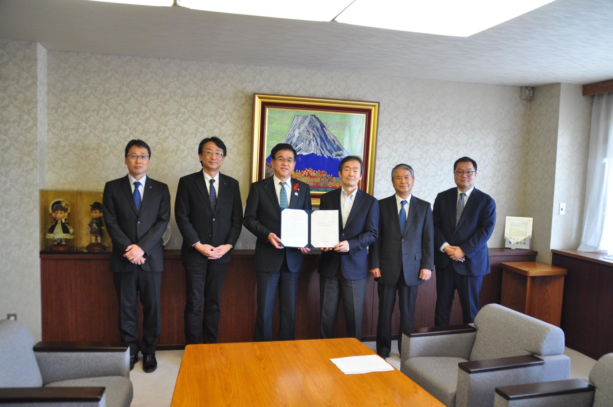 スーツ姿の男性6人が室内で並び、中央の市長と板野部会長が書類を手にして記念撮影をしている様子の写
