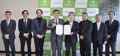 白い紙を広げて持つスーツ姿の市長と板野会長を中心に、両側にそれぞれ3人ずつ合計8人の男性が直立して並び、緑と白の「HADANO秦野市」のロゴが背景にある集合写真