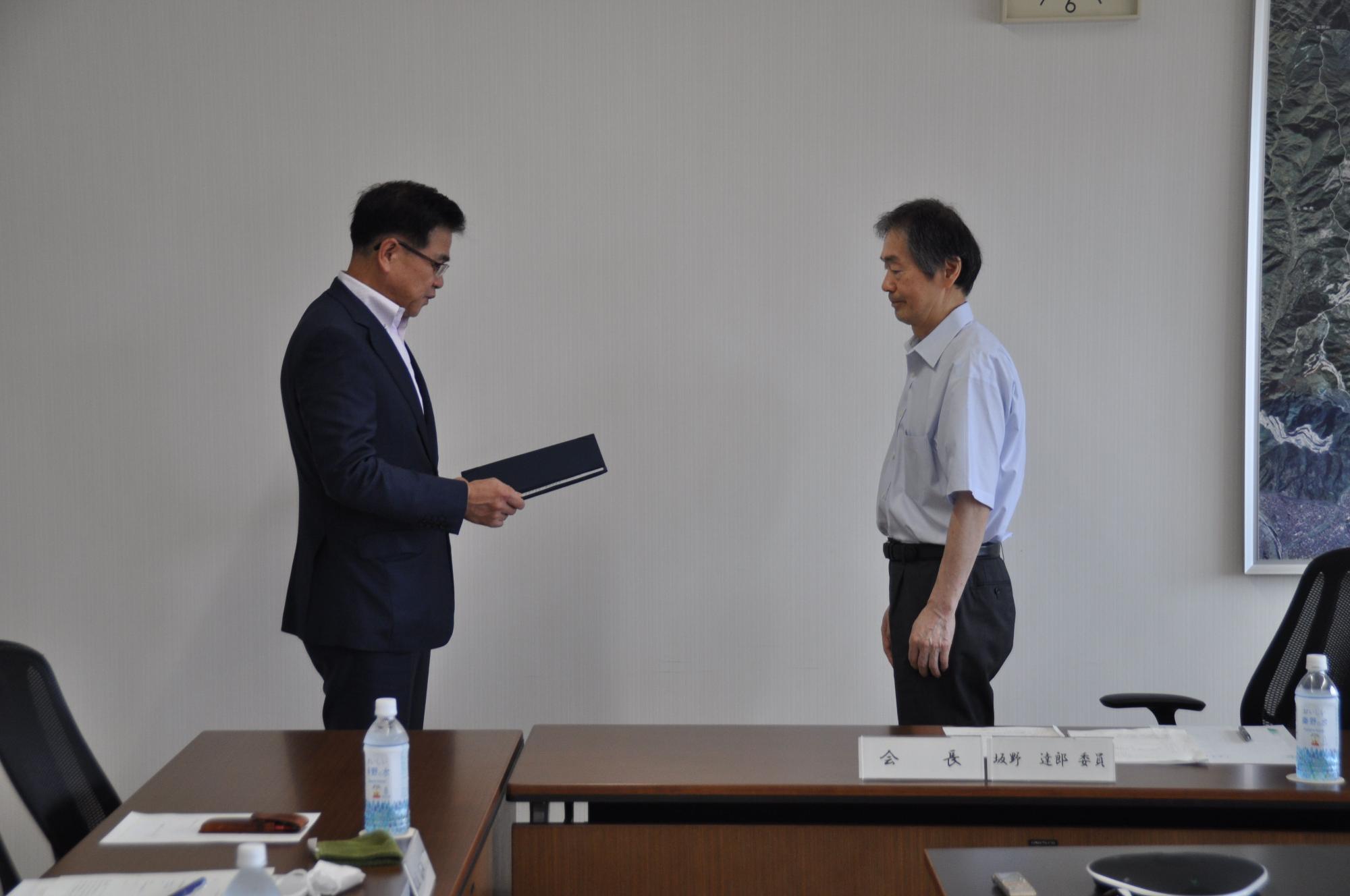 白い壁の会議室で、紺色のスーツ姿の市長が黒いファイルを持ち、白シャツ姿の板野会長と向かい合って書類授受を行っている場面の写真
