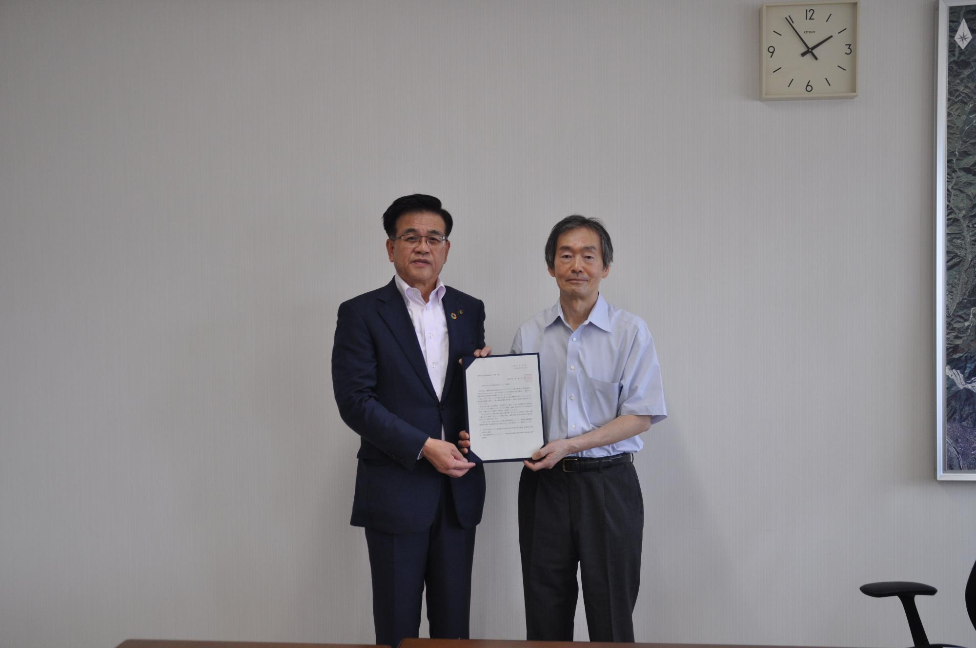 白い壁と時計が掛かった室内で、紺色のスーツ姿の市長と水色のシャツ姿の板野会長が並んで諮問書を掲げ、記念撮影している写真