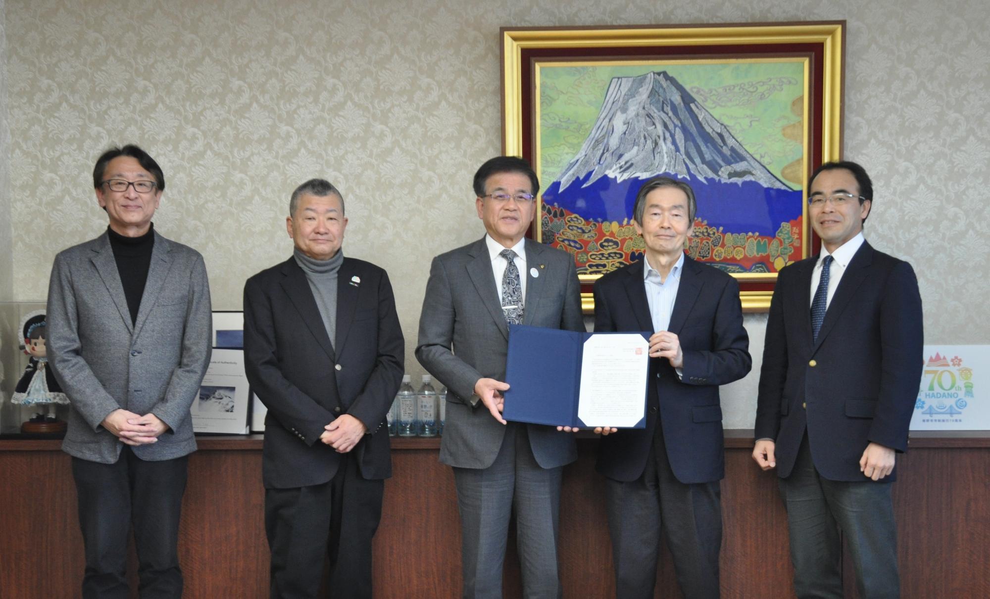 金色の額縁に入った富士山の絵画を背景に、ビジネスカジュアルからスーツ姿の5名の男性が横一列に並び、中央の市長と板野会長報告書を掲げる公式行事の写真