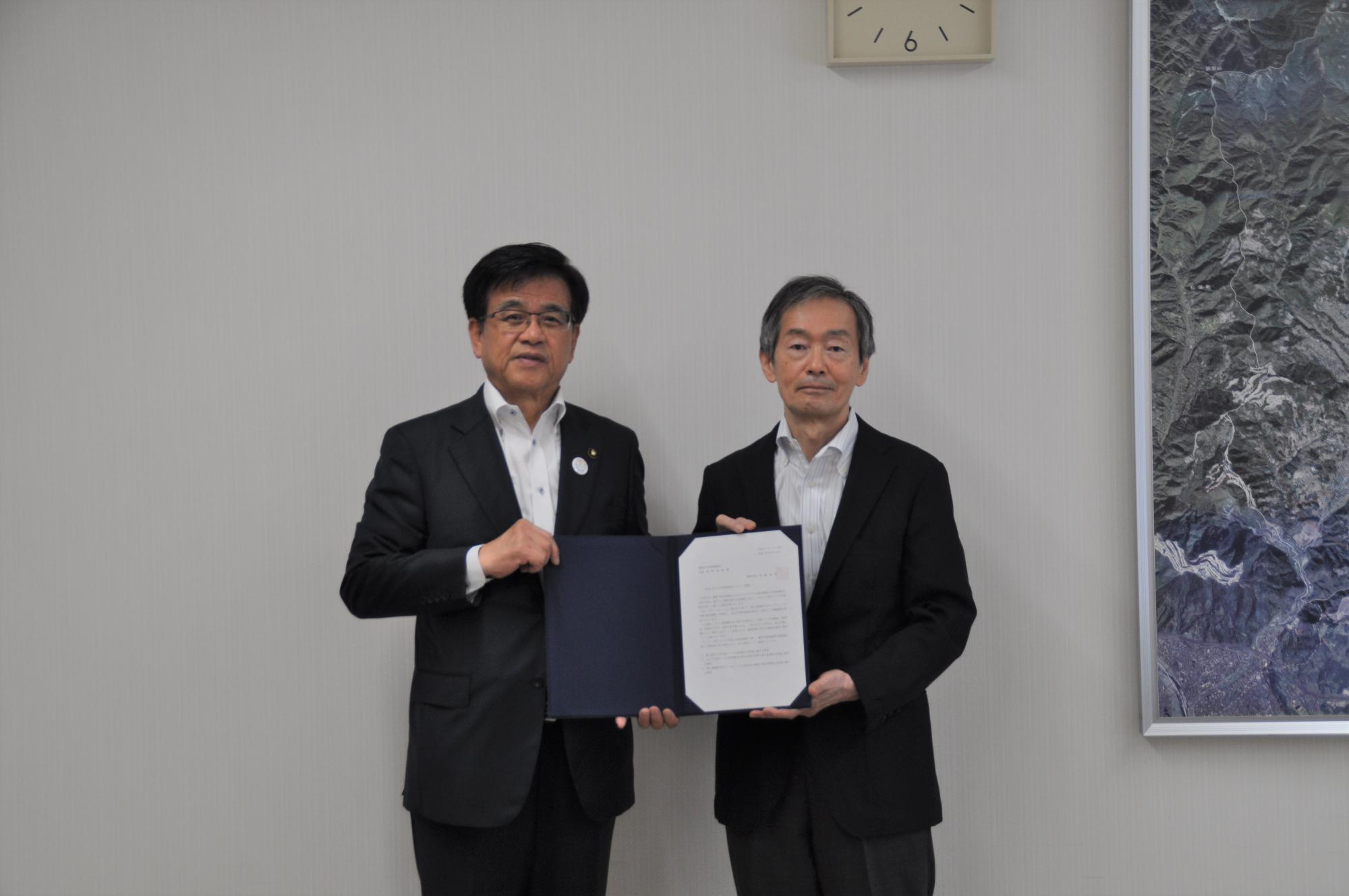 スーツを着た市長と板野会長が、一緒にネイビーのフォルダーに挟まれた書類をカメラに向かって提示している様子の写真