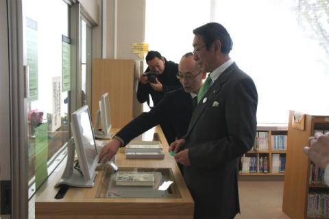 図書室でスーツを着た二人の男性がモニターが付いた機器が並んだ木製のカウンター前に立ち、その背後から男性がカメラを構えている様子の写真