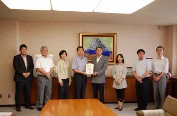 応接室らしき場所で、スーツ姿やカジュアルな服装の8人が富士山のような絵画の下で書類の受け渡しをしている様子の記念写真