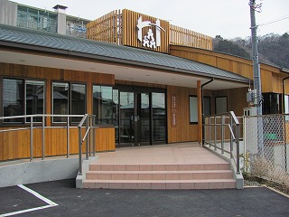 木目調の外壁と濃い色の屋根で、正面玄関前の階段やスロープに手すりが設置されている、障がい者地域活動支援センターの外観写真