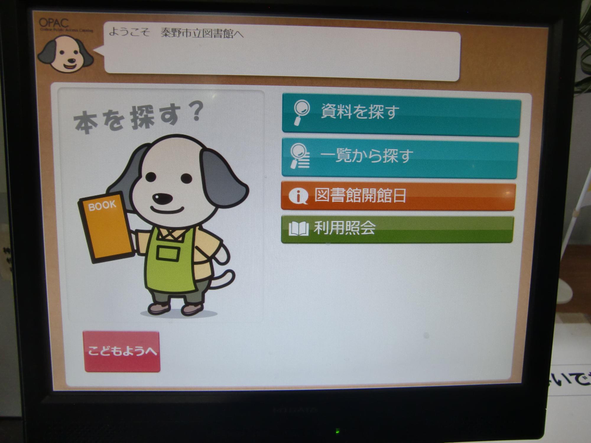 画面に表示された犬のキャラクターが本を抱え、その横に「資料を探す」「一覧から探す」などのメニューが並ぶ秦野市立図書館のOPAC画面の写真