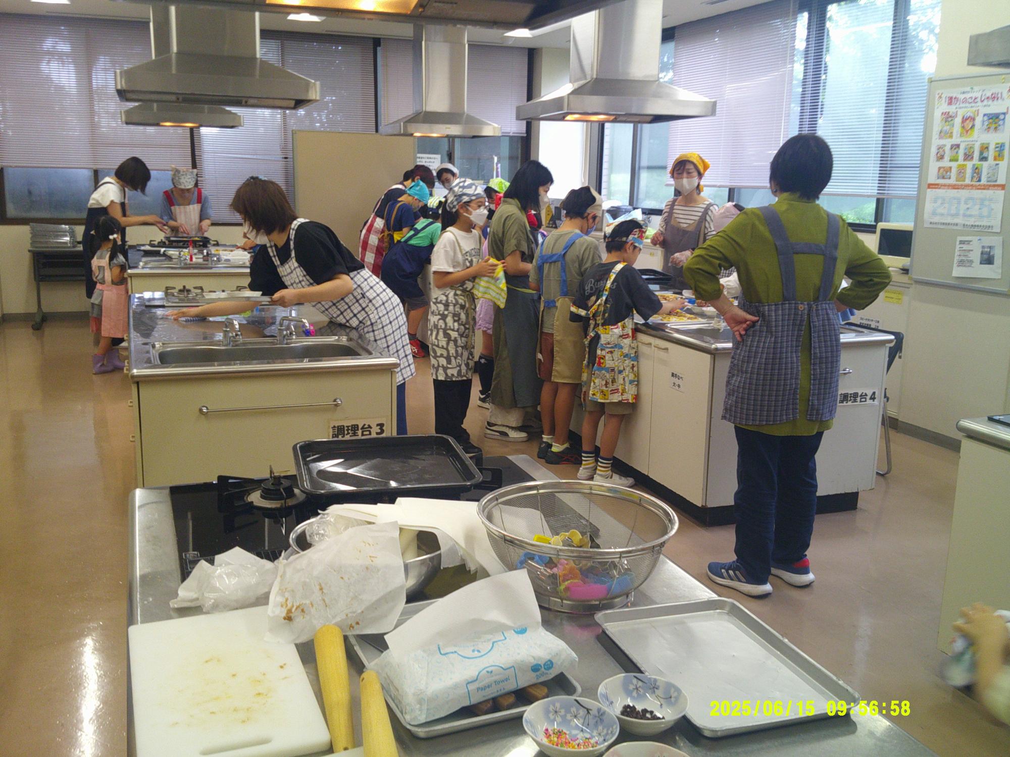 複数の調理台と換気扇が設置された清潔な実習室で、エプロンを着用した大人と子供たちが料理や調理の活動に熱心に取り組んでいる写真