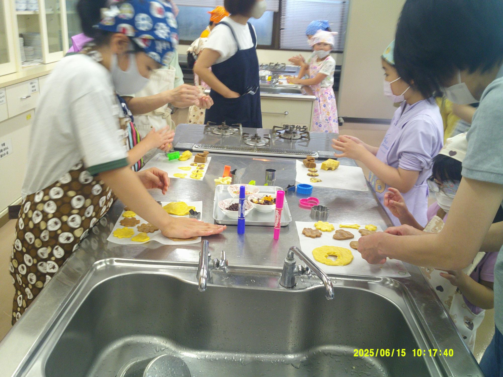 調理台を囲んで大人と子供たちがマスクと三角巾を着用し、クッキーのような生地を型抜きしたり飾り付けたりしている様子の写真