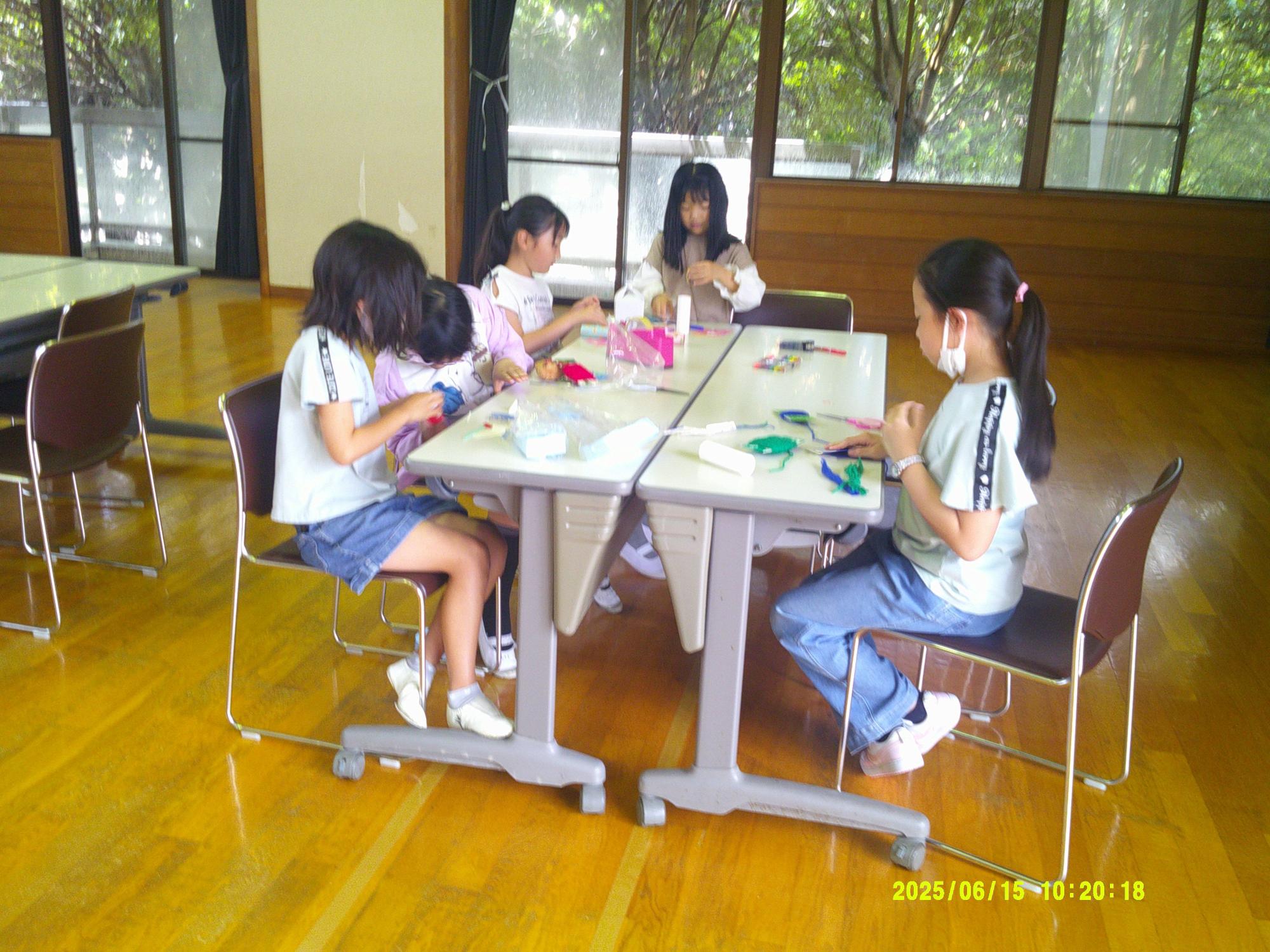 木目調の床の明るい室内で、数人の子供たちが二つ連結された机を囲んで座り、ミニ工作活動に集中して取り組んでいる写真