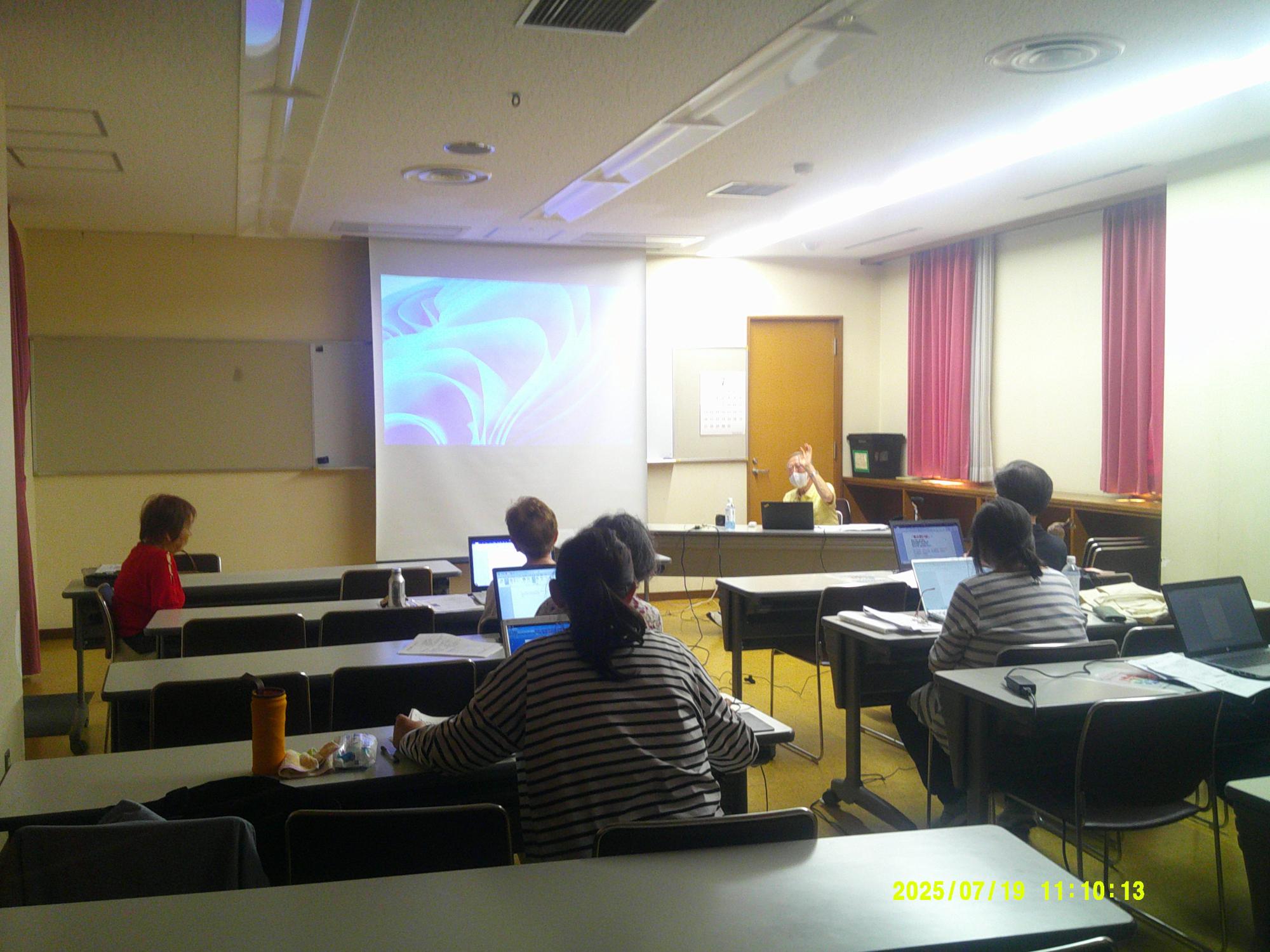会議室のような一室で、参加者がパソコンを机の上で開き席に座っており、講師が中央のスクリーンに画面を投影しながら説明をしている様子の写真