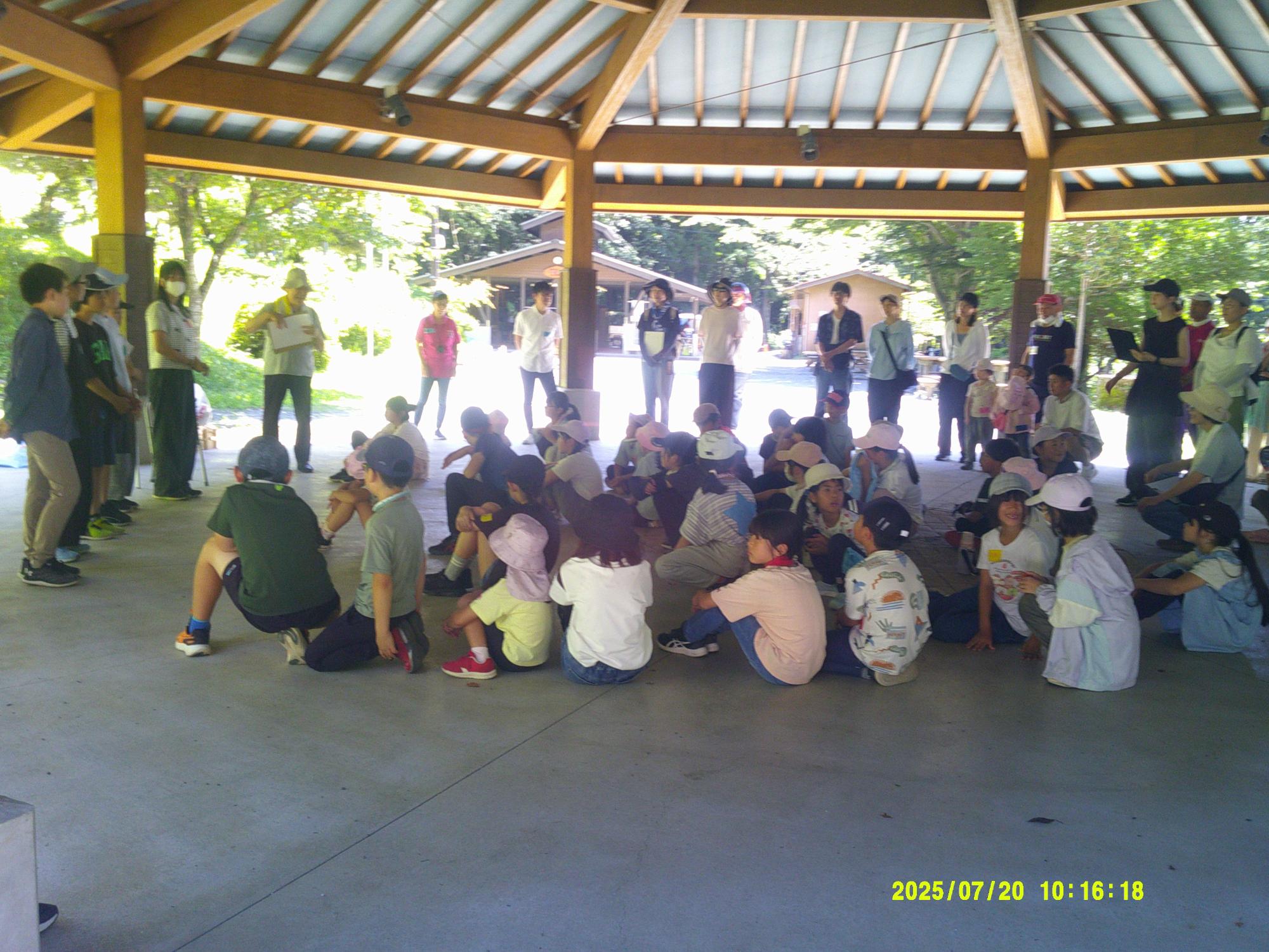 大きな屋根の下の休憩所で、大勢の子供たちが地面に座り、数人の大人が立っている様子を捉えており、自然に囲まれた屋外での写真
