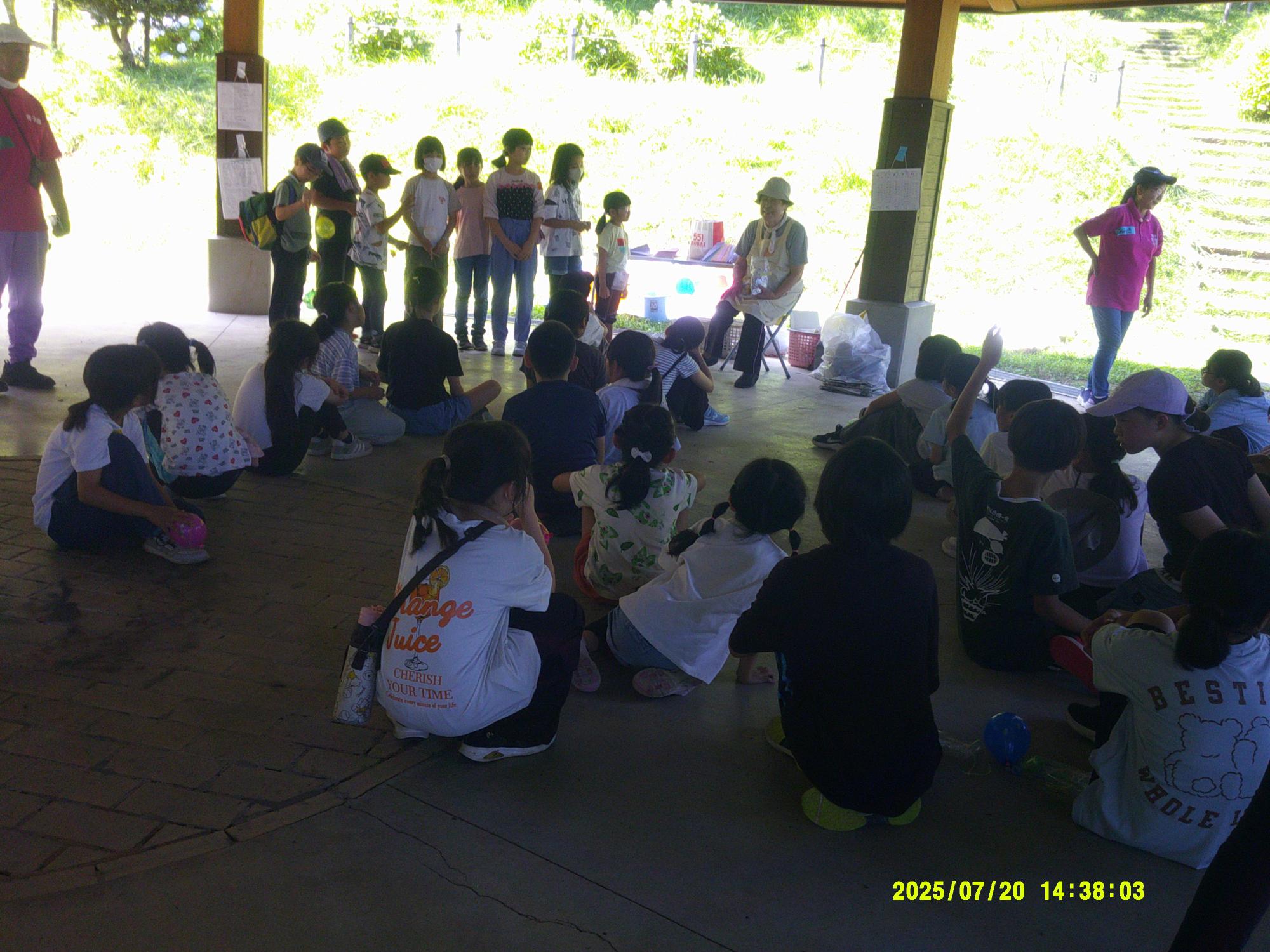 屋外の屋根付きの休憩所で、大勢の子供たちが地面に座って説明を聞いており、数人の大人が立っている様子の写真