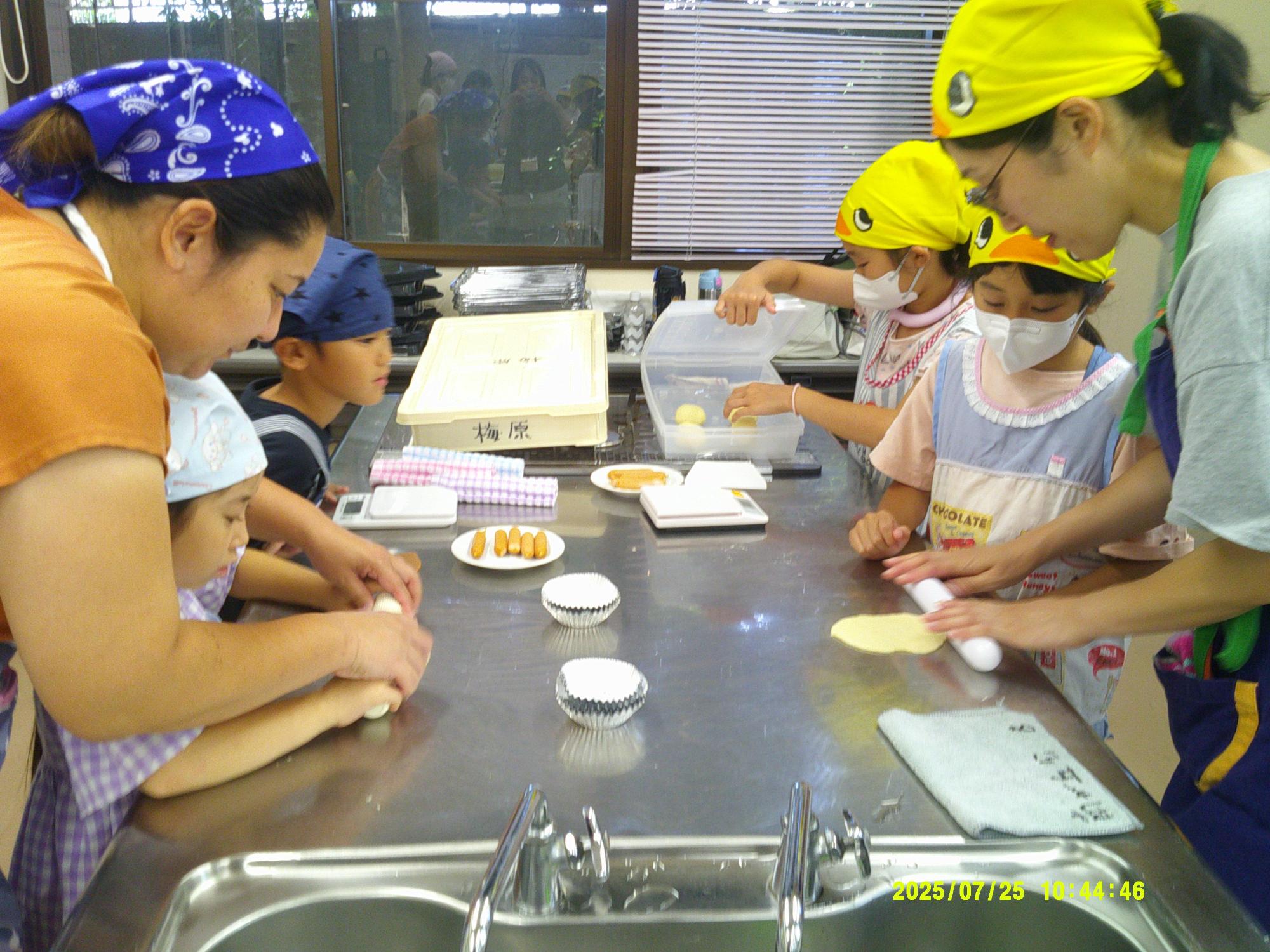 調理場で、三角巾とマスクをつけた親子連れが、調理台の上でパン生地を伸ばしたりこねたりしている写真