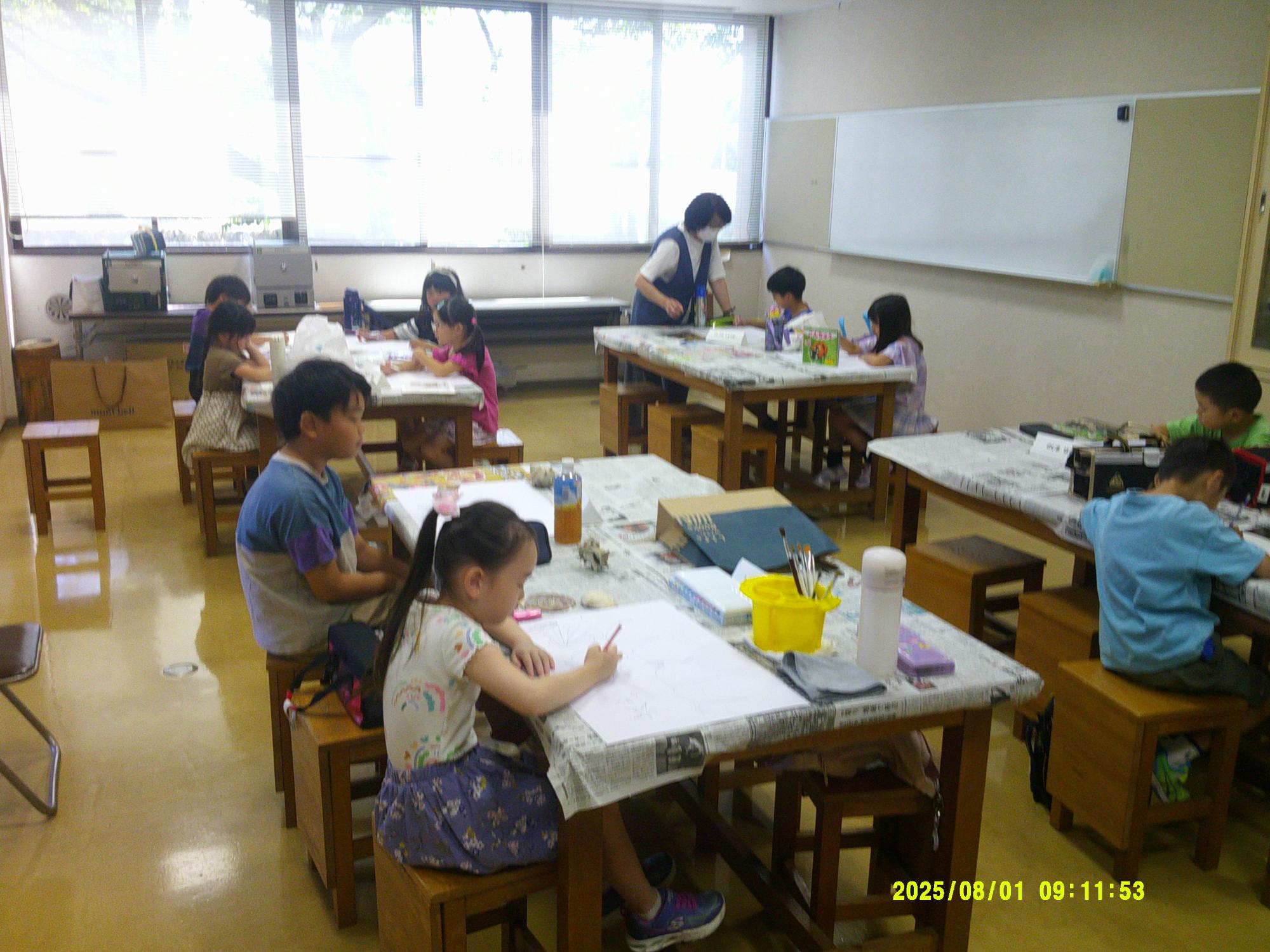 大きな窓のある教室で、数人の子どもたちが机に向かい絵を描いたり工作をしたりしており、奥ではエプロン姿の先生が指導にあたっている絵画教室の様子の写真