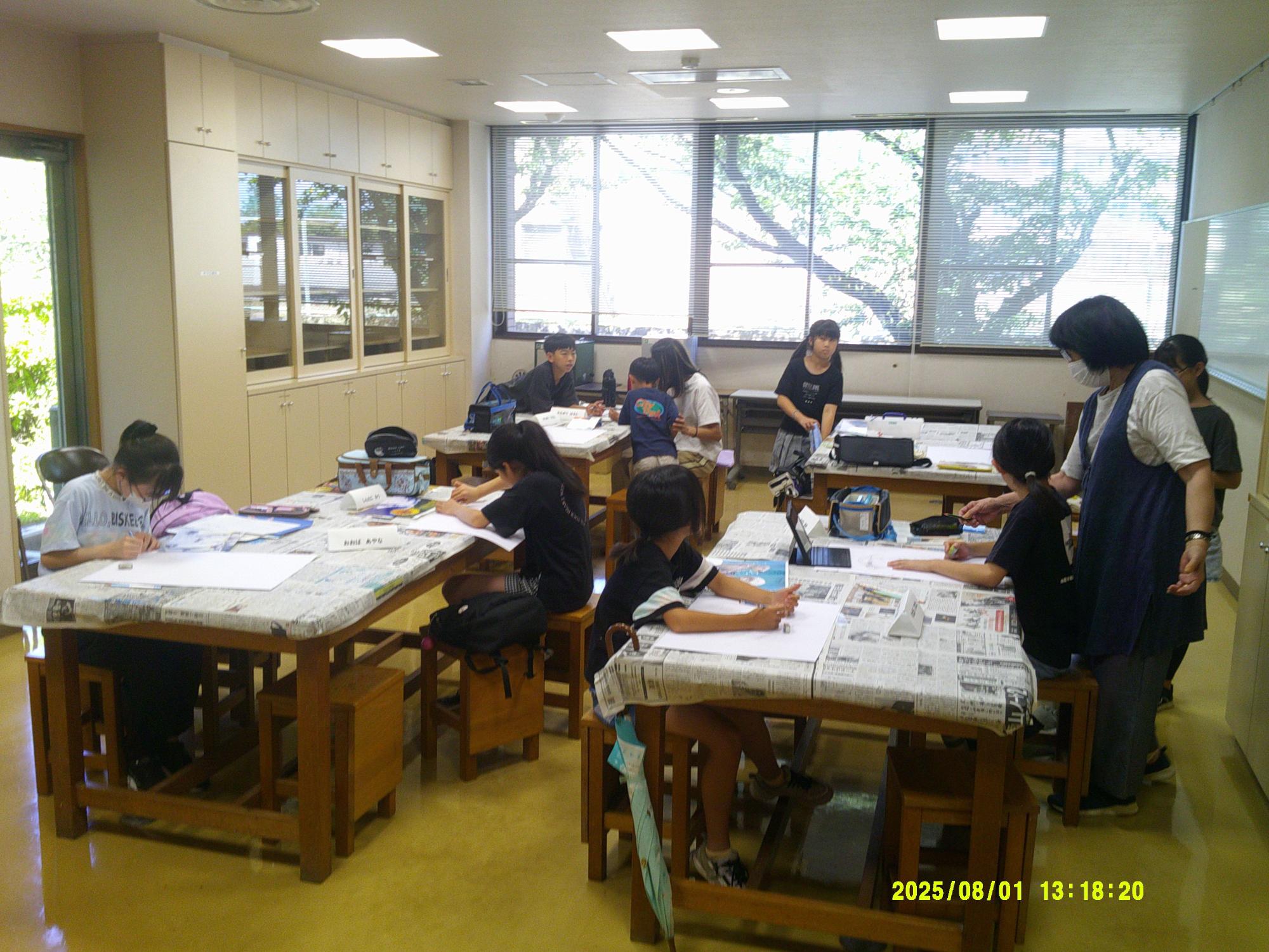 大きな窓のある教室で、数名の小学生が机に広げた新聞紙の上で、鉛筆や絵の具を使い熱心に制作活動に取り組み、先生らしき女性が立って指導している絵画教室の写真
