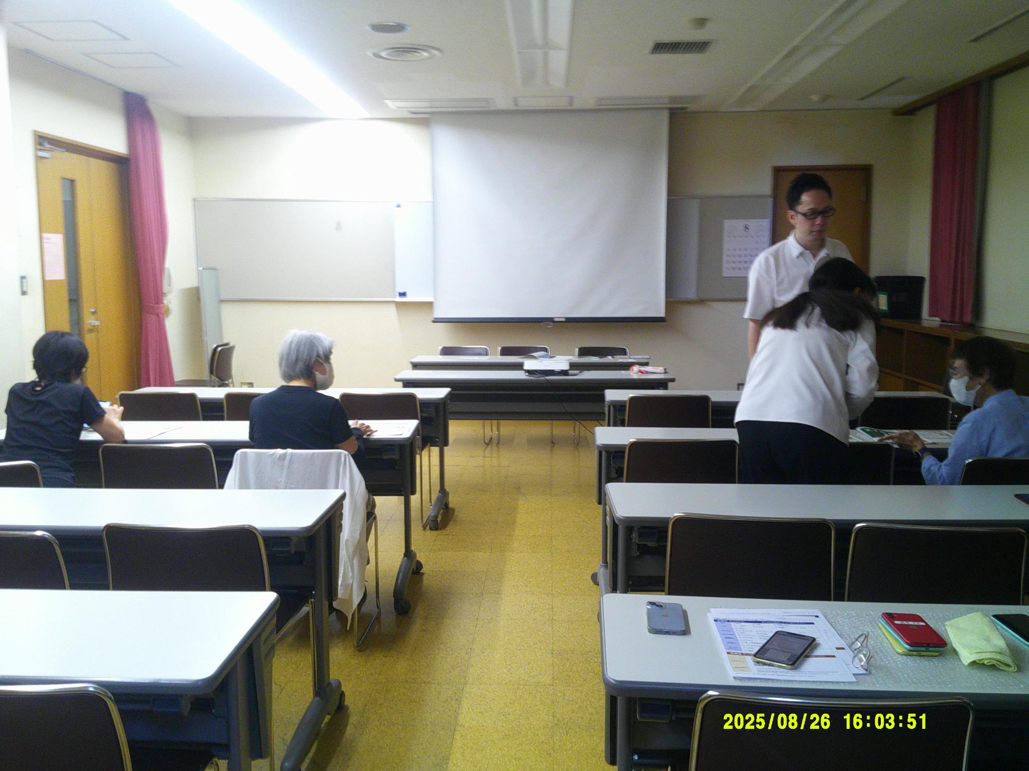 広いスクリーンとホワイトボードのある室内で、数名の参加者が席に座っており、講師らしき男性が説明している様子の写真