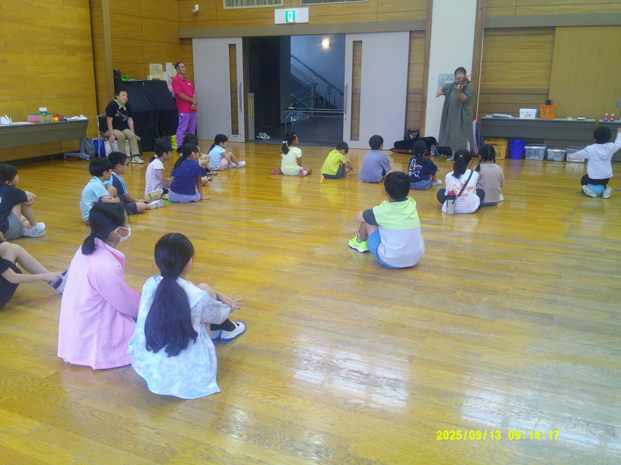 木目調の広い室内で、多くの子どもたちが床に座り、奥の大人たちの方向を向いて説明を聞いている様子を捉えた写真