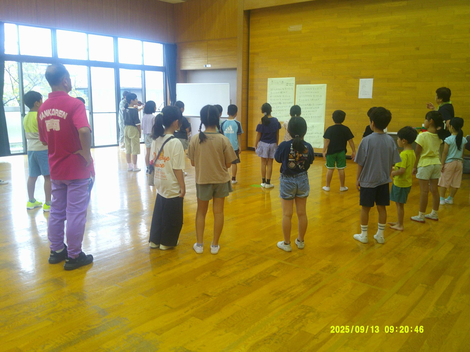 黄色い木目調の床と大きな窓がある明るい場所で、多数の子供たちが大人の指導員を囲むようにして立っている様子を後ろから写した写真